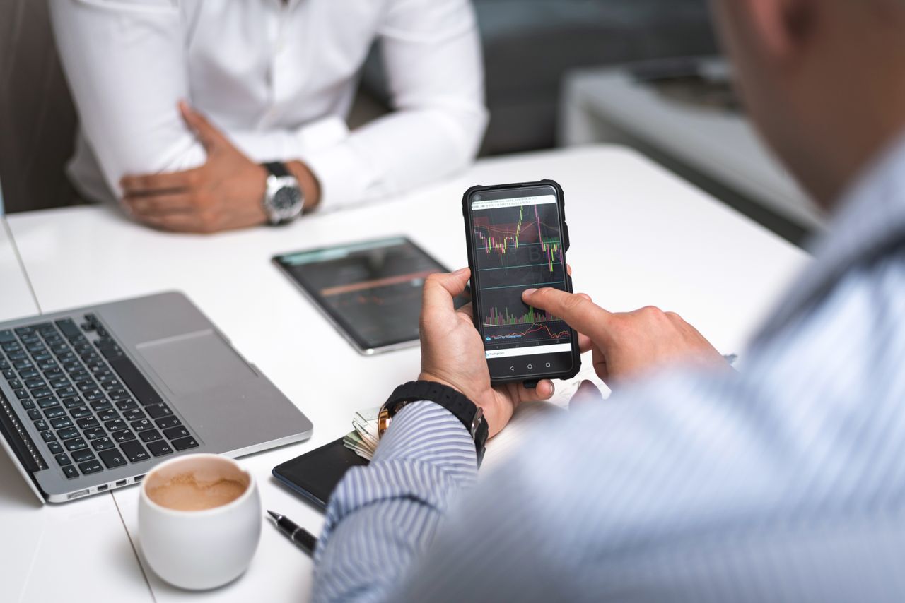 Two businessmen analyze stock market data using a smartphone and laptop in a modern office setting.